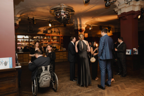 Attendees in formal attire stand and talk in a reception space with a bar and lounge seating, one attendee seated in a wheelchair with a backpack nearby