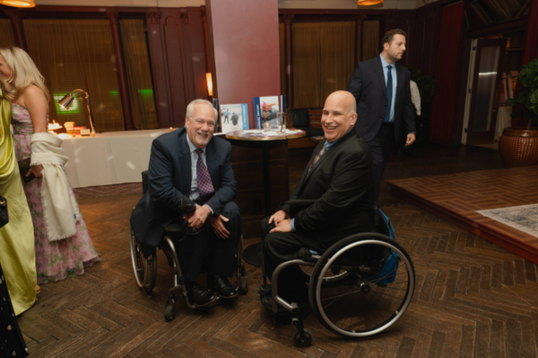 Two attendees in suits seated in wheelchairs at a cocktail table during a CAF reception with event materials displayed behind them
