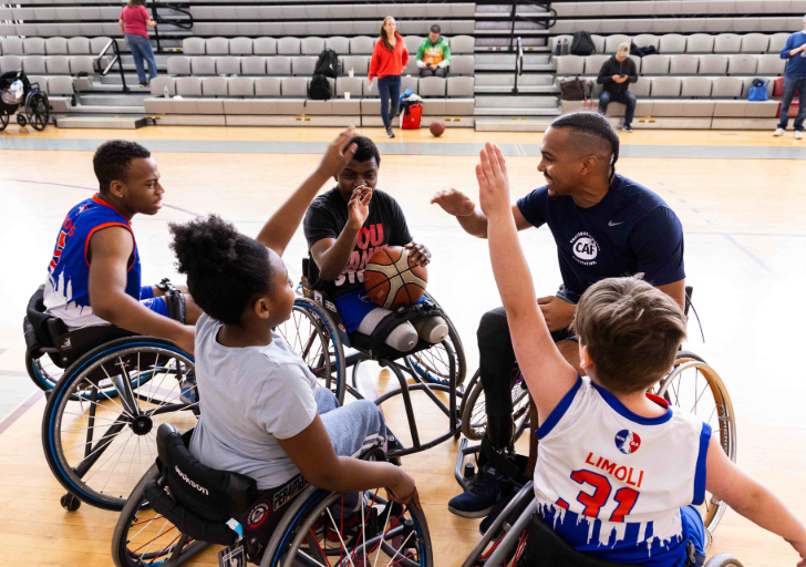 Participants using sports wheelchairs form a circle on a gym court, raising hands toward a group high-five with a basketball held in the center