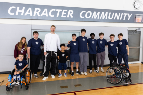 Youth volunteers and participants lined up along a gym wall beneath the words Character Community, with a sports wheelchair positioned at the side