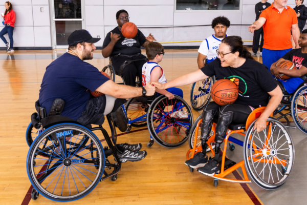 Participants in sports wheelchairs on a gym court exchanging a fist bump while holding basketballs, with others positioned around the group