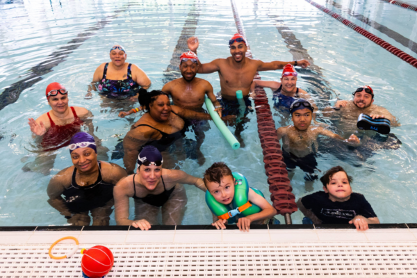 Participants gathered in a swimming pool during a clinic activity, positioned along a lane line with flotation devices and pool equipment visible