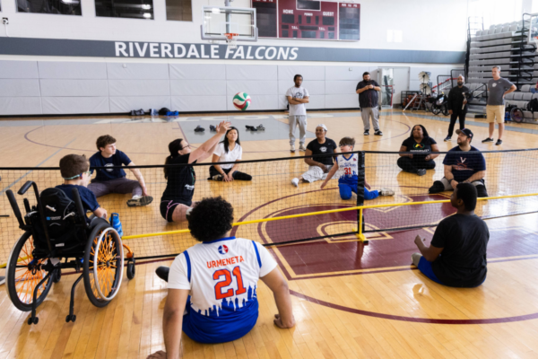 Participants seated on a gym floor around a low net, passing a ball across the court during a seated volleyball activity with Riverdale Falcons signage on the wall