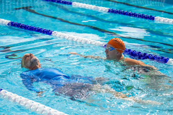 Two people in a lap lane, one floating on their back while another faces them in the water with lane lines on either side.