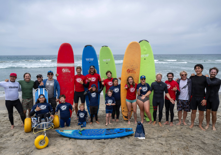 Group of people wearing CAF shirts standing on a beach with surfboards arranged vertically in the sand, ocean waves in the background, adaptive beach wheelchair and surfboard in the foreground