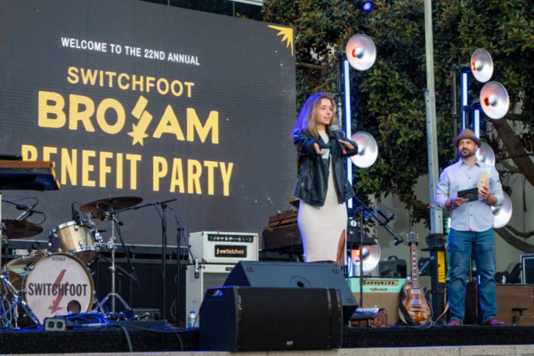 Stage setup with drum kit, guitars, and speakers beside a large screen reading Welcome to the 22nd Annual Switchfoot BroAm Benefit Party, two speakers on stage holding microphones
