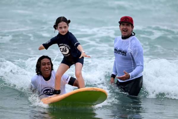 Participant standing on a surfboard in the water with two instructors nearby providing support, small waves breaking around them