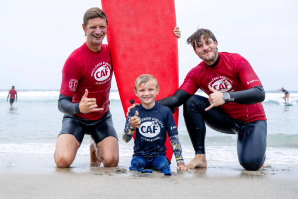 Two instructors in red CAF shirts kneeling beside a participant sitting on the sand with a red surfboard positioned upright behind them, ocean in the background