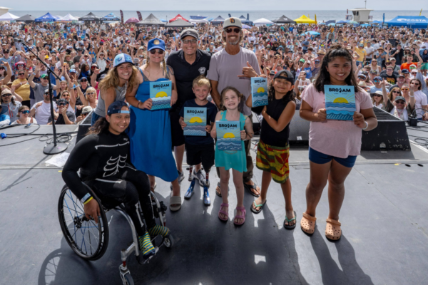 Group of participants and supporters on stage holding BroAm posters with a large crowd gathered on the beach behind them, tents and sponsor booths visible along the shoreline