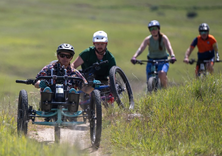 A group of cyclists on a grassy terrain, with one using an adaptive three-wheeled cycle in the foreground.