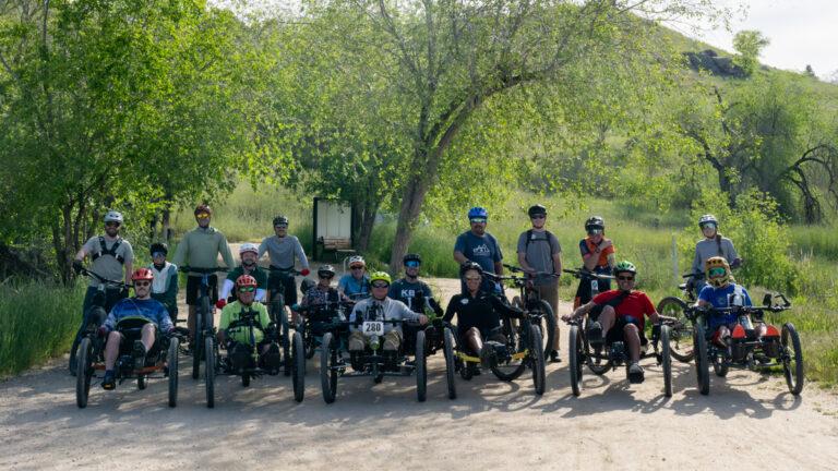 A group of people with all-terrain tricycles, wearing helmets, in a green outdoor setting with trees and grass.