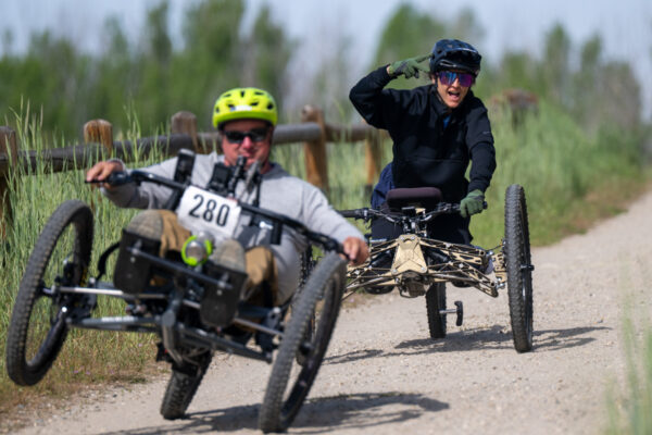 Two people riding adaptive bicycles on a gravel path with a wooden fence and greenery in the background.