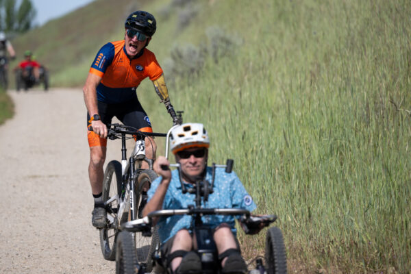 Cyclists on a dirt path with the foreground showing a man in a blue shirt on a handcycle, followed by a man on a bicycle wearing an orange outfit.