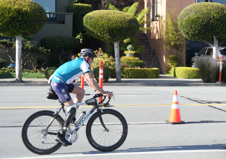 Cyclist riding a road bike along a city street marked with orange traffic cones, wearing a helmet and a cycling kit, with a residential neighborhood and parked cars in the background