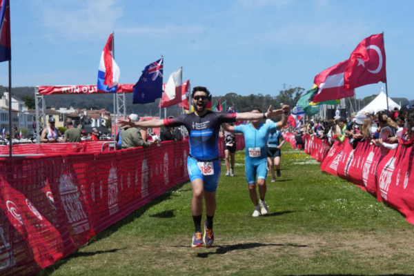 Athlete running across a grassy finish chute lined with red barriers and international flags, arms outstretched, with other runners approaching behind
