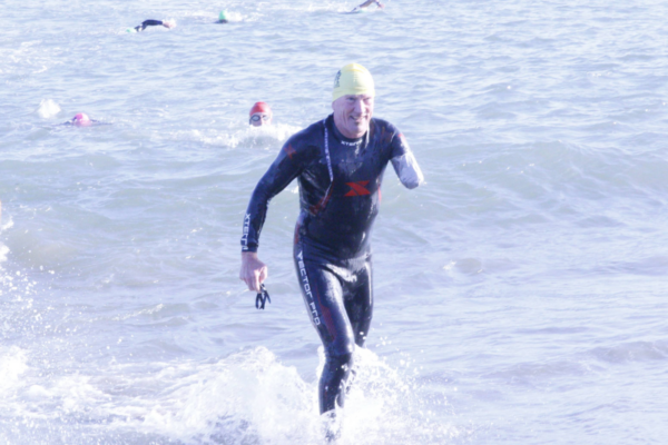 Athlete in a wetsuit and swim cap stepping out of open water onto shore holding goggles, with additional swimmers visible in the water behind