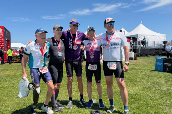 Five athletes wearing medals standing together on grass after a race, with event tents and signage visible in the background