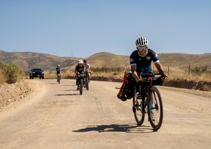 Cyclists ride along a rural dirt road during a long-distance ride, with rolling hills and support vehicles in the background.