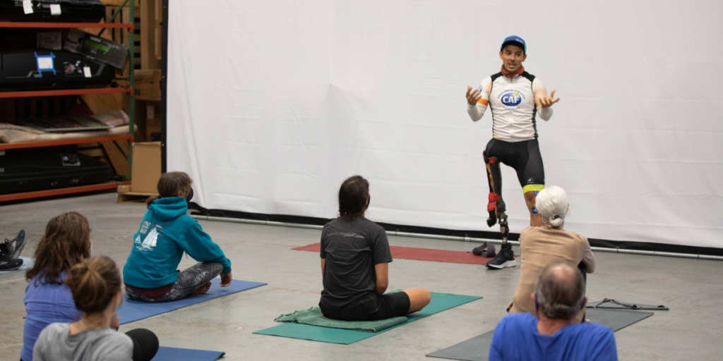 Athlete with a prosthetic leg leads a small group through a seated stretching or mobility session indoors.