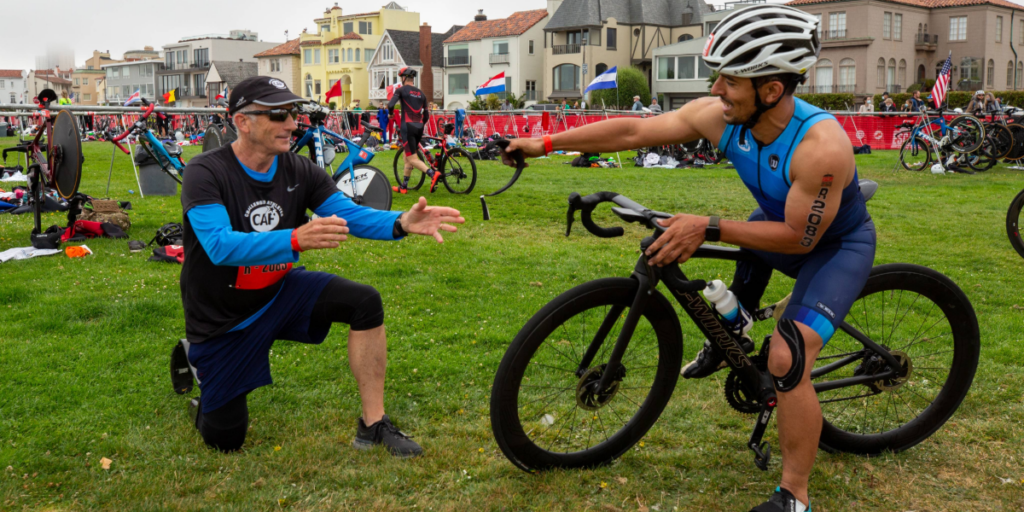 Triathlete on a bike reaches out to a supporter in a transition area lined with gear and bikes along a coastal course.