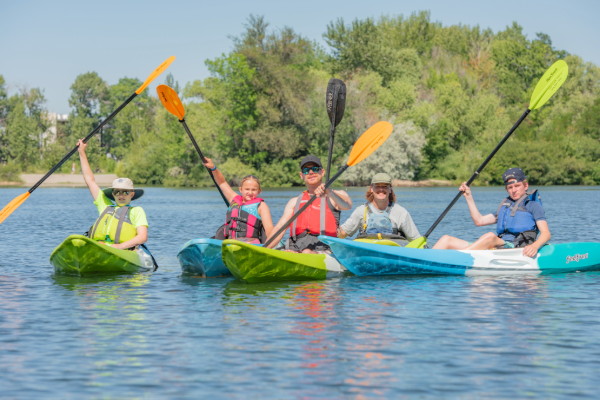 Participants paddle colorful kayaks across calm water, raising paddles in the air, with trees lining the shoreline in the background.
