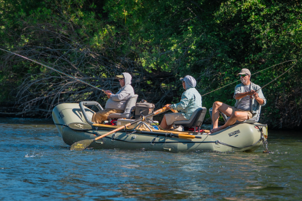 Three participants sit in an inflatable raft on a river, each holding a fishing rod, surrounded by dense green vegetation along the bank.
