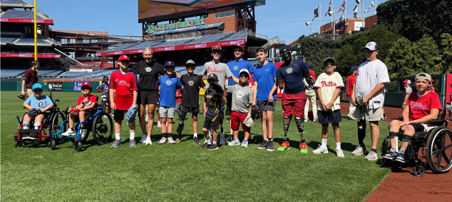 Group of youth participants and coaches standing on a baseball field at Citizens Bank Park during the Phillies Coaches Clinic, including several athletes using wheelchairs, with the stadium scoreboard and seating in the background.