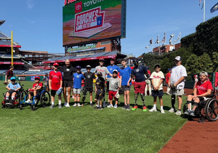 Group of youth participants and coaches standing on a baseball field at Citizens Bank Park during the Phillies Coaches Clinic, including several athletes using wheelchairs, with the stadium scoreboard and seating in the background.