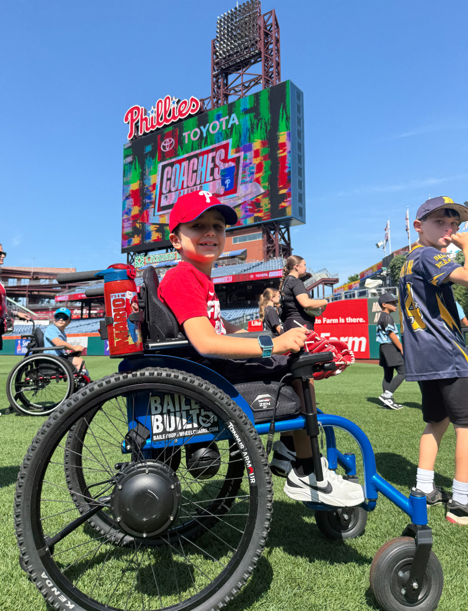 Young athlete using a blue adaptive sports wheelchair on the field at Citizens Bank Park, holding a baseball glove, with other participants and the Phillies Coaches Clinic scoreboard visible behind.