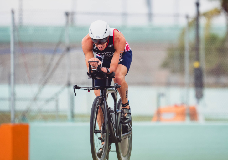 Triathlete in an aerodynamic position rides a time trial bike on a paved course, wearing a helmet and tri suit, with barriers and fencing in the background.
