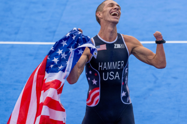 Athlete holds a U.S. flag while standing on a blue track surface, wearing a triathlon suit with USA lettering.