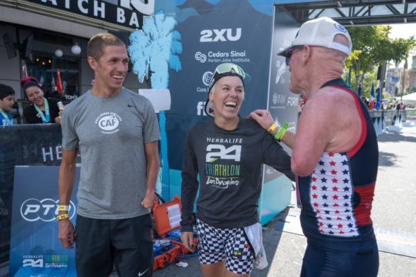 Three athletes stand and talk near a triathlon event area with sponsor signage and equipment visible behind them.