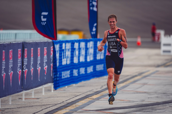 Triathlete runs along a race course lined with banners and barriers, wearing a racing kit and moving toward the finish area.
