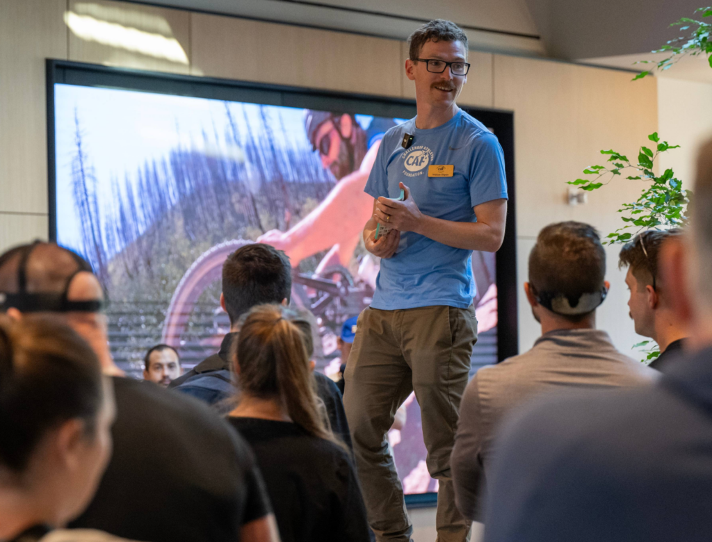 A presenter speaks to a group of symposium attendees during a CAF Idaho educational session, with adaptive sport imagery displayed on a screen behind.