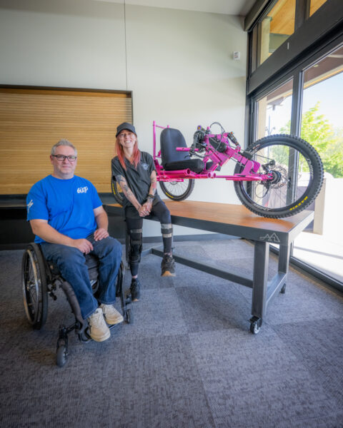 Two individuals in a room with an adaptive pink handcycle on a table.