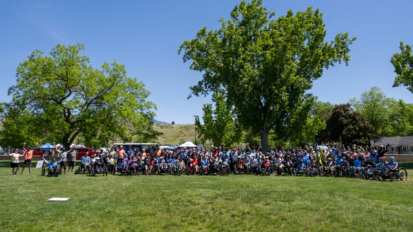 A large group of people, many in wheelchairs or bikes, gathered in a park under blue skies with trees and hills in the background.