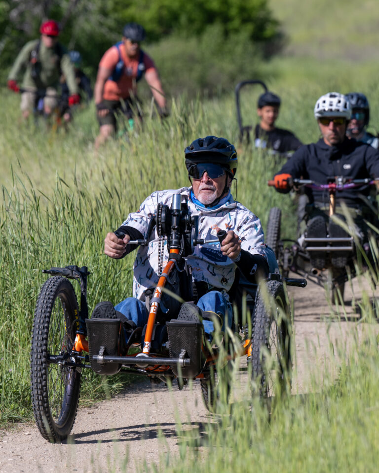 A group of cyclists on recumbent tricycles ride through a grassy trail.