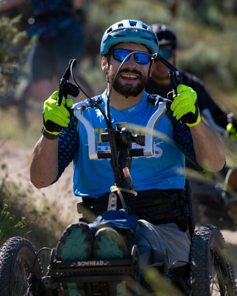 A person riding an adaptive mountain bike with a bright blue helmet and neon gloves on a dirt trail.