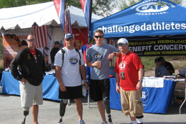 Athletes with lower limb amputations stand in front of a Challenged Athletes Foundation event booth during a community adaptive sports gathering.