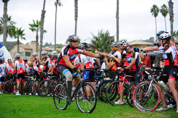 An athlete with a lower limb amputation rides a road bike and reaches toward fellow participants during a Challenged Athletes Foundation cycling event.