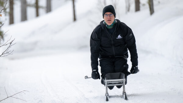 A person wearing black winter clothing is sitting on a sled, navigating through a snowy forest path.