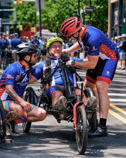 Three men assist a smiling individual in a handcycle during a cycling event. One man crouches beside him while another adjusts the cycle.