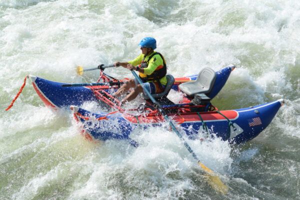 A person in a yellow life jacket and blue helmet paddles an inflatable blue raft through whitewater rapids.