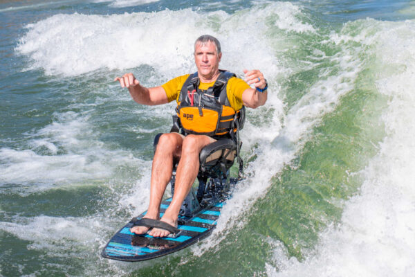 A man with short gray hair smiles while riding an adaptive wakeboard on water, dressed in a yellow shirt and a black life jacket, with splashing waves around him.