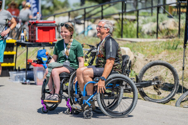 Two individuals in wheelchairs sit on a paved path, smiling in a sunny outdoor setting surrounded by bicycles and outdoor equipment.