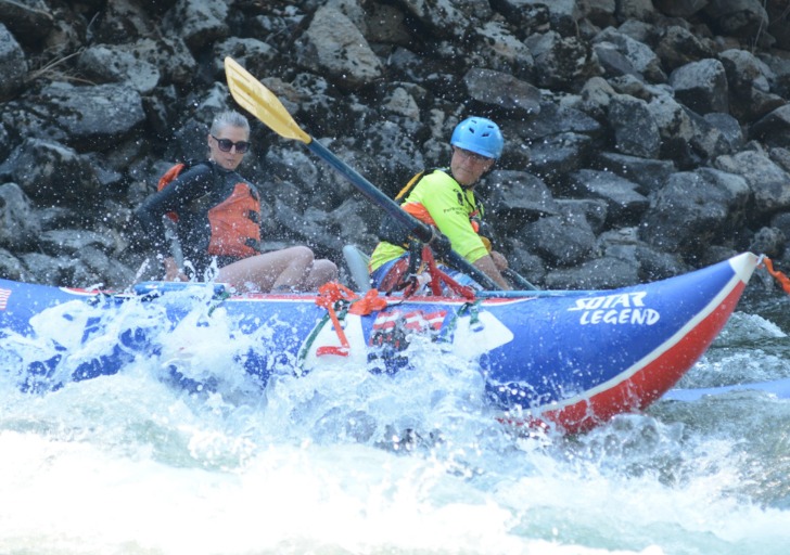 Two individuals in a colorful raft labeled "SOLAR LEGEND" navigate choppy river waters; one woman in an orange life jacket and sunglasses, and a man in a blue helmet and yellow shirt.