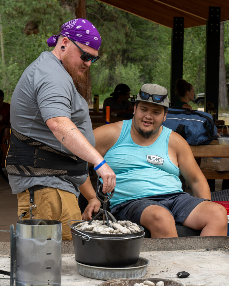 Two men outdoors engaged in cooking; one is using tongs to handle a pot, while the other observes.