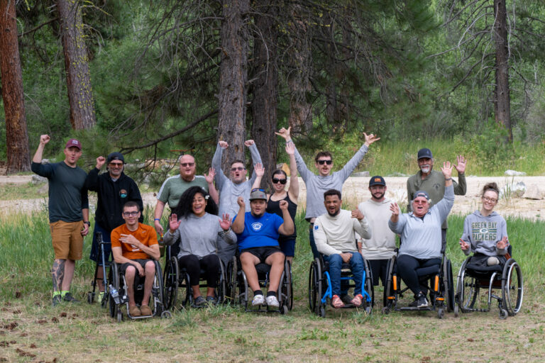 A diverse group of fourteen people, some in wheelchairs, poses outdoors, smiling and raising their arms in celebration against a backdrop of trees and grass.
