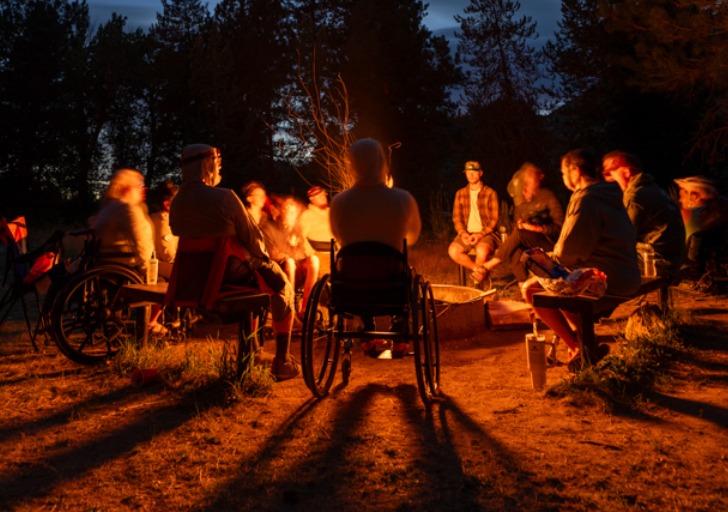 A diverse group of people, some in wheelchairs, sit around a campfire during dusk, illuminated by the fire's orange glow, with trees and a twilight sky in the background.