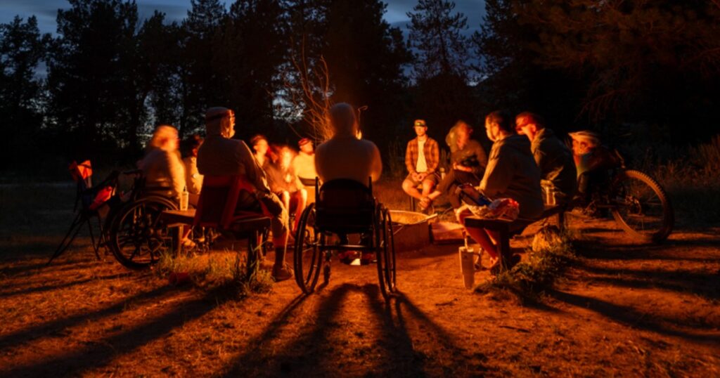 A diverse group of people, some in wheelchairs, sit around a campfire during dusk, illuminated by the fire's orange glow, with trees and a twilight sky in the background.